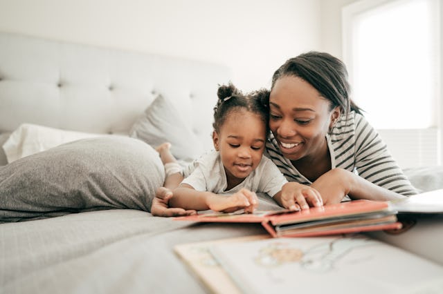 A mother and her young daughter lie on a bed, happily reading a book together, enjoying a cozy and e...