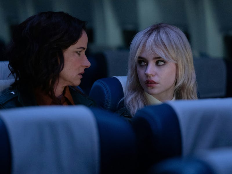 Two women sit close together in an airplane cabin, engaged in an intense conversation. The dim light...