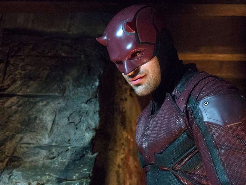 A man dressed in a red superhero costume with a mask stands in a dimly lit basement. He has a determ...