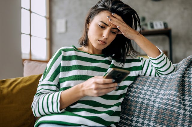 A young woman sits on a couch, wearing a green and white striped sweater, with a concerned expressio...