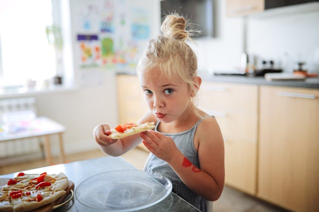 A young girl with blonde hair tied in a bun sits at a kitchen table, eating a slice of pizza topped ...