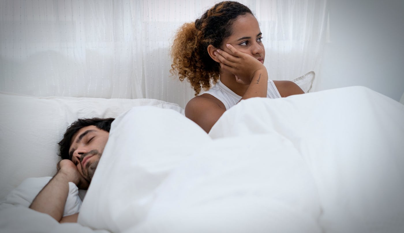 A woman sits upright in bed with a thoughtful expression, while a man sleeps beside her. They are covered with white blankets in a softly lit room.