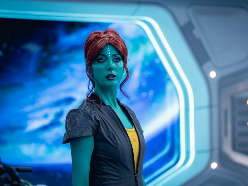 A woman with blue skin and red hair stands in a futuristic environment, featuring glowing panels and...
