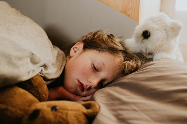 A child lies peacefully asleep in bed, covered by a blanket, with a teddy bear and a plush polar bea...