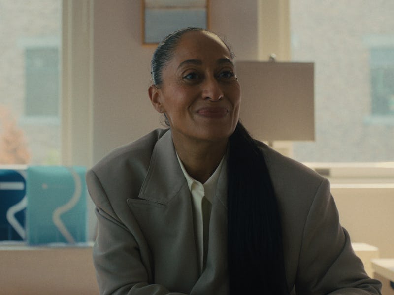 A smiling woman in a tailored gray suit sits at a desk in a bright office with large windows. She ha...