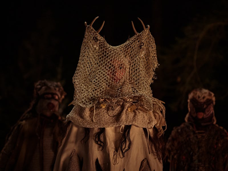 Three figures in the dark woods, dressed in eerie costumes with netting and animal-like masks. A sur...