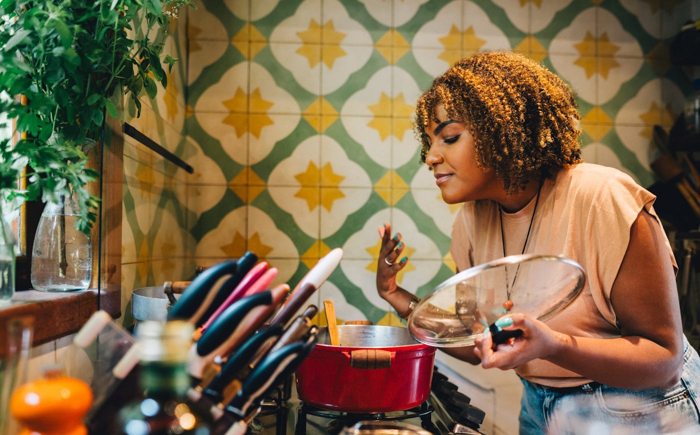 A young woman with curly hair joyfully cooks in a cozy kitchen, holding a lid over a red pot, surrounded by colorful tiles and cooking utensils.