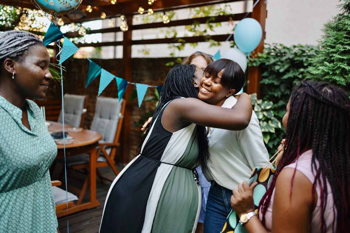 A joyful gathering of women celebrating, with one woman embracing another. The outdoor space is decorated with balloons and colorful bunting.