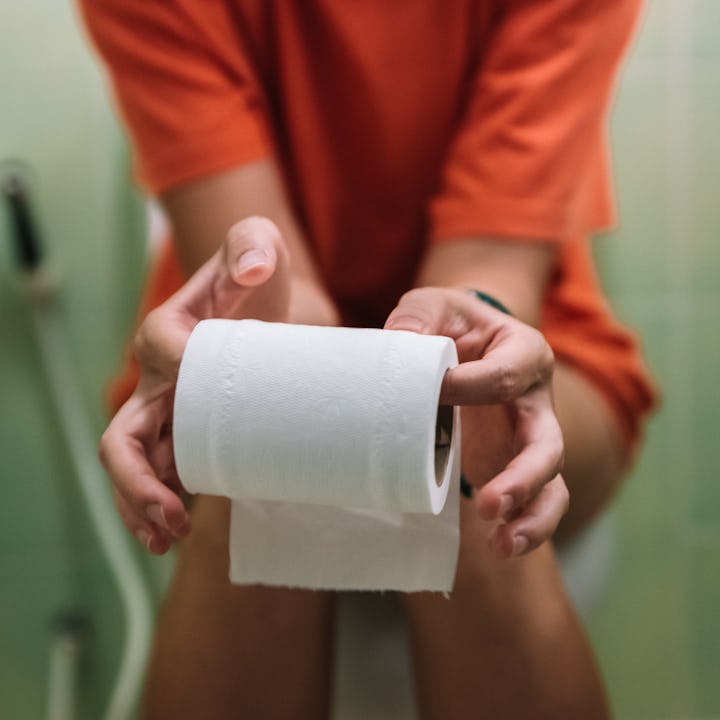 A person in an orange shirt holds a roll of toilet paper with both hands, seated on a toilet. The ba...