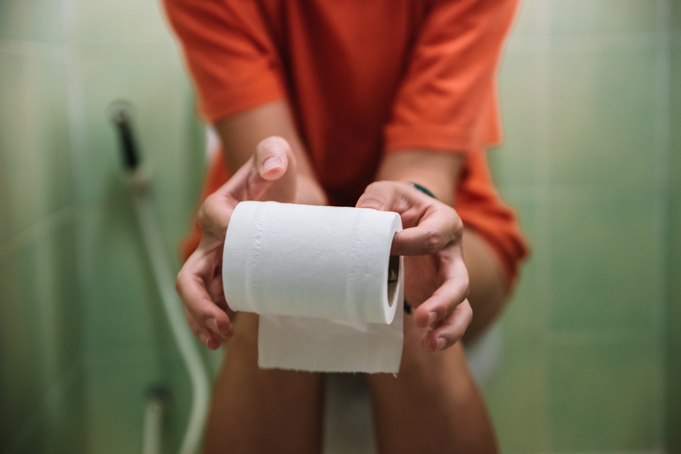 A person in an orange shirt holds a roll of toilet paper with both hands, seated on a toilet. The bathroom has green tiled walls in the background.