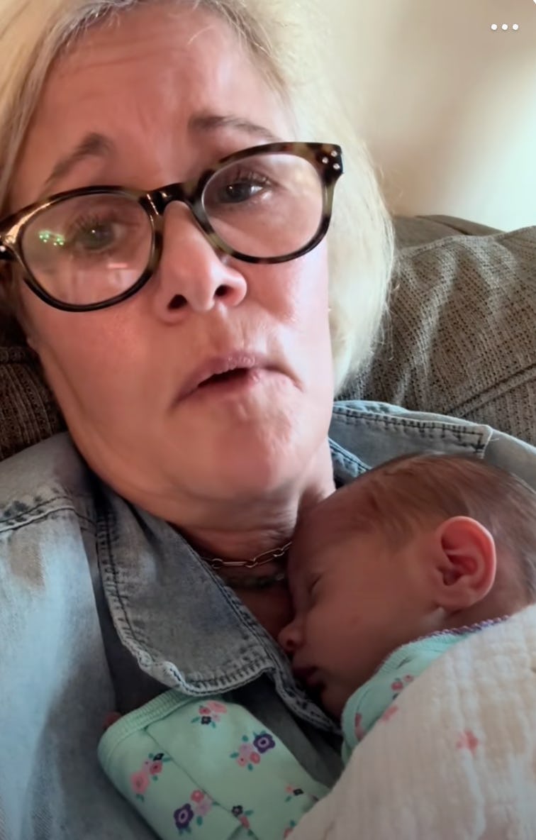 A woman with glasses looks directly at the camera while holding a sleeping baby close to her chest, both resting on a cozy couch.