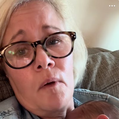 A woman with glasses looks directly at the camera while holding a sleeping baby close to her chest, both resting on a cozy couch.