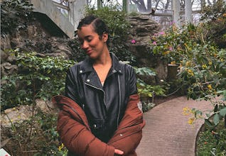 A young woman with curly hair stands in a lush garden setting, wearing a black leather jacket and a …