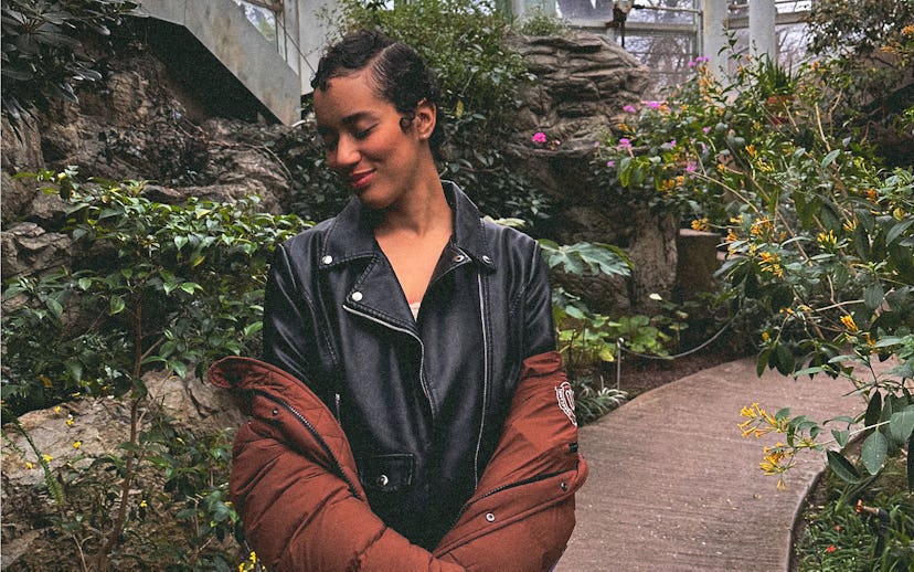A young woman with curly hair stands in a lush garden setting, wearing a black leather jacket and a ...