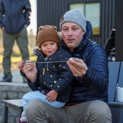 A man sits on a chair, holding a stick with a child next to him, both smiling. In the background, another person stands near a fire pit.