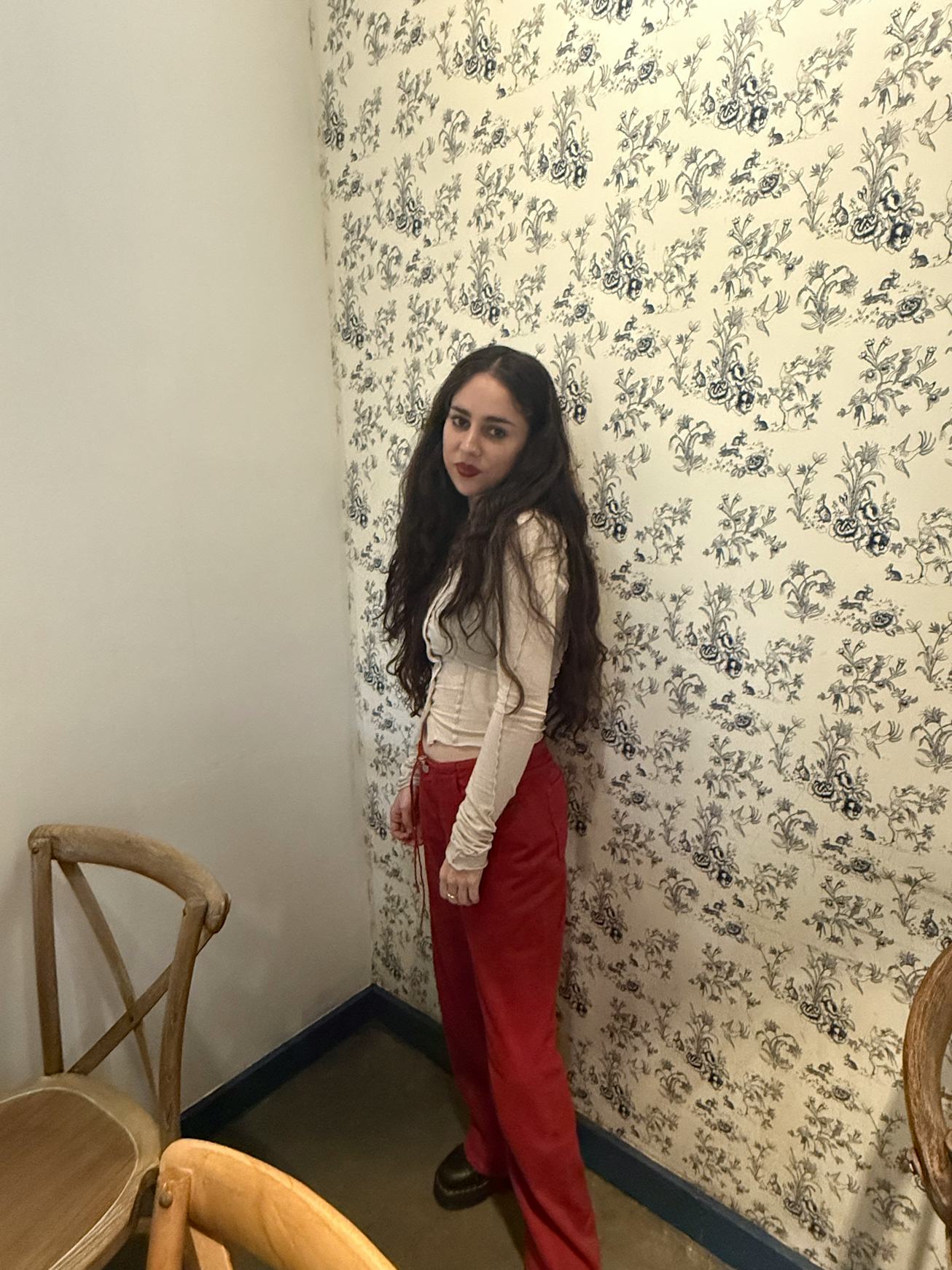 A young woman with long, dark hair stands against a patterned wall. She wears a light top and red pa...