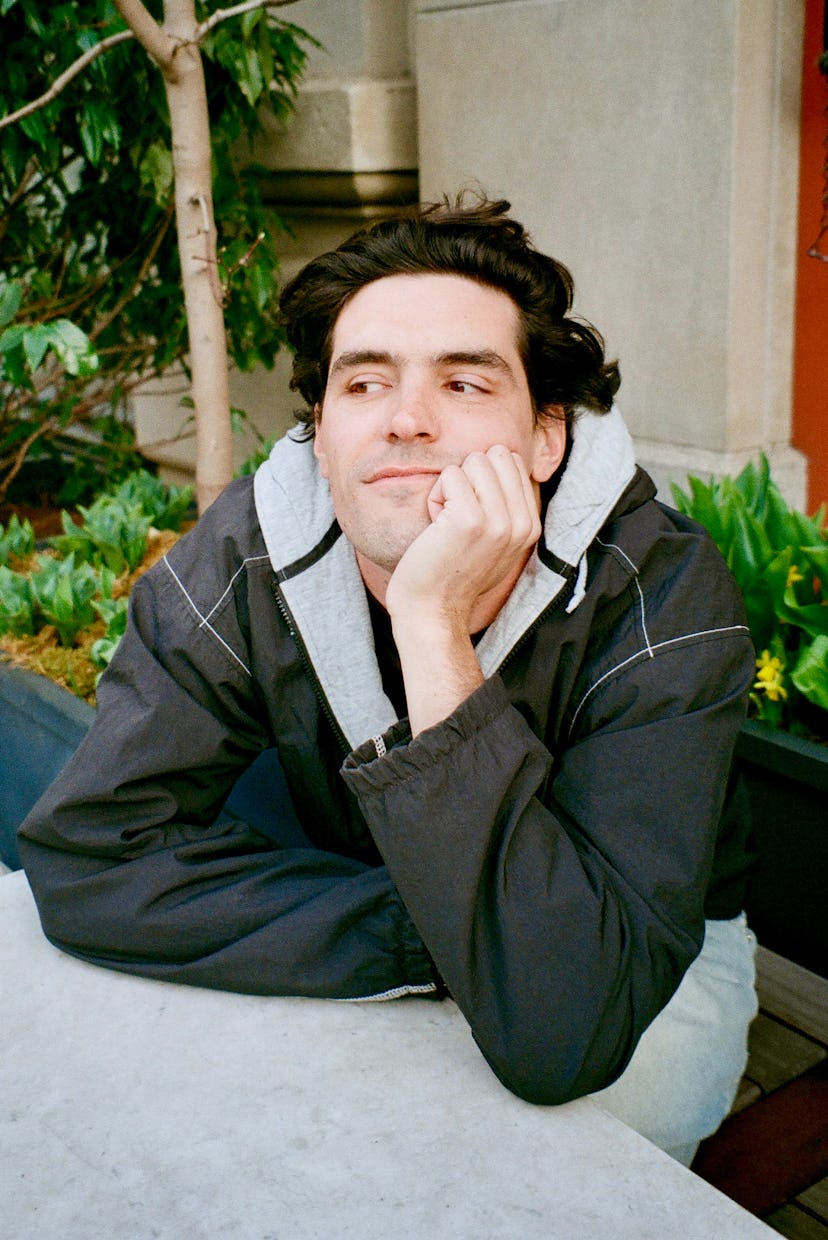 A casually dressed young man rests his chin on his hand while sitting at a table outdoors, surrounde...