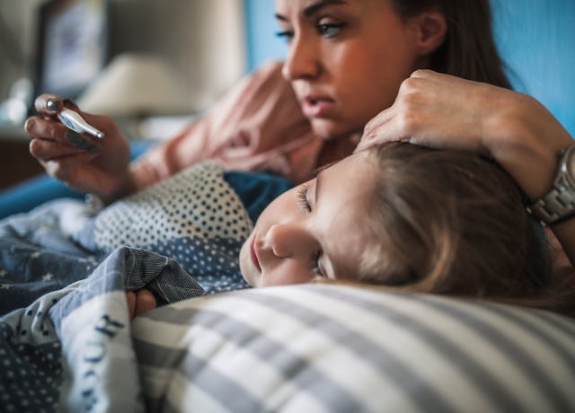 A woman gently holds a child's head while looking at a phone, both resting on a bed with patterned p...