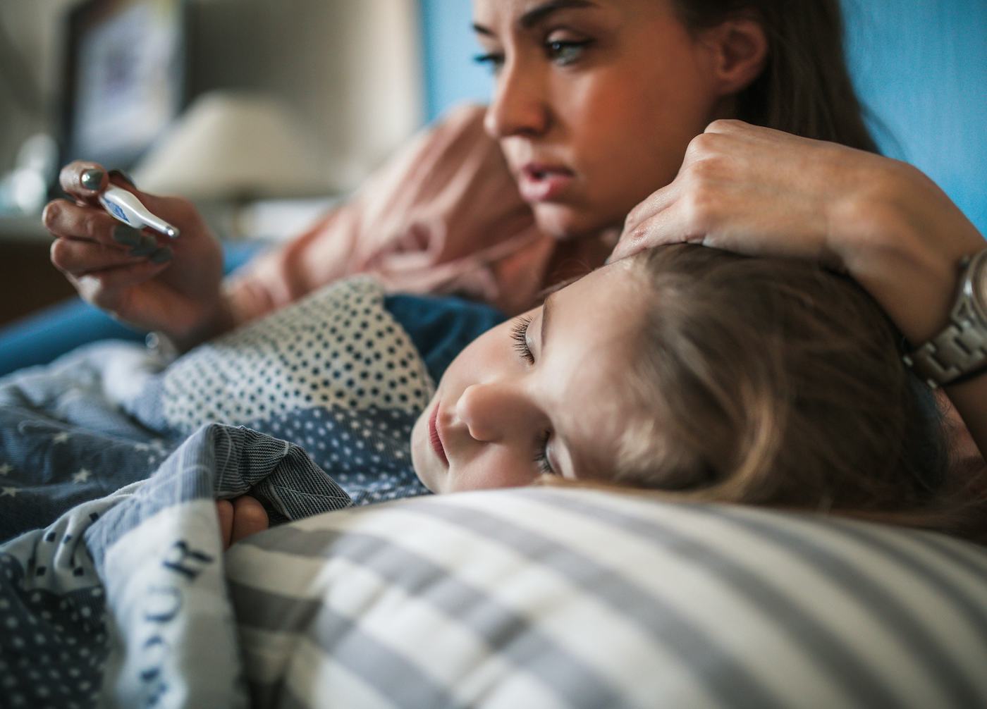 A woman gently holds a child's head while looking at a phone, both resting on a bed with patterned pillows, conveying a tender, caring moment.