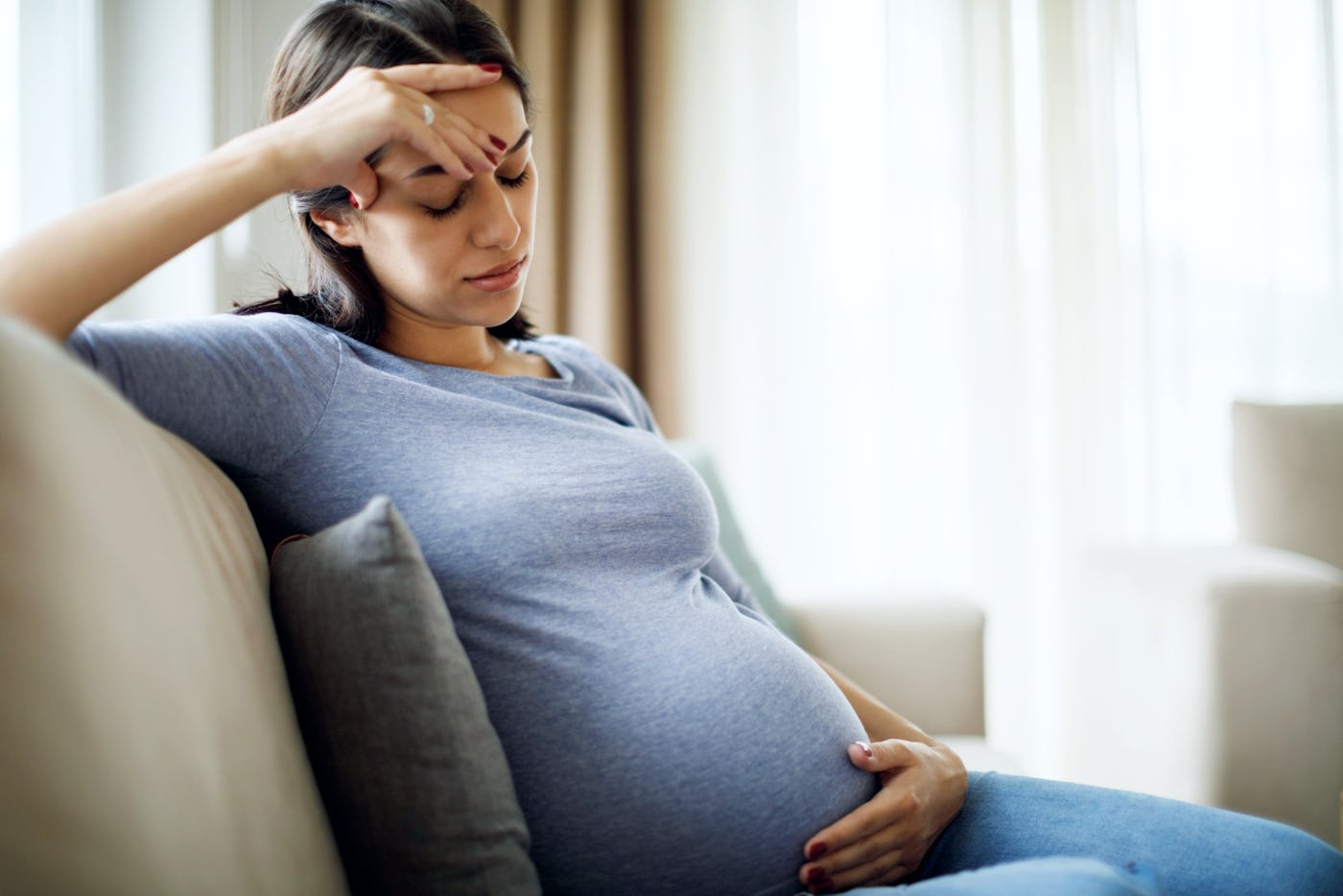 A pregnant woman sits on a couch, resting her forehead on her hand. She looks contemplative while gently cradling her baby bump. Natural light filters in.