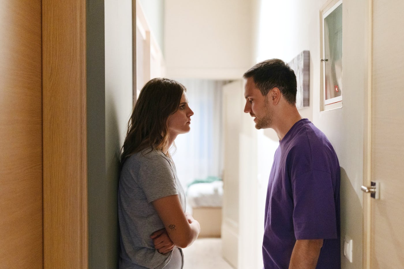 A woman in a gray shirt and a man in a purple shirt stand facing each other in a hallway, both displaying intense expressions, suggesting a serious conversation.