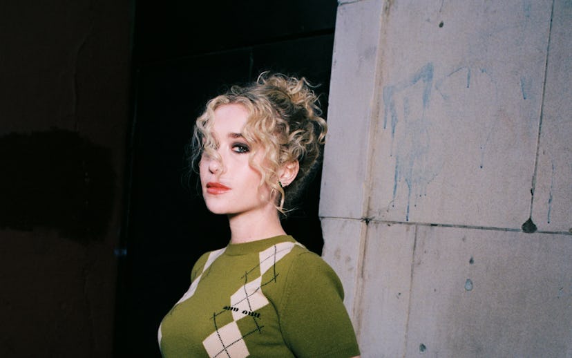 A young person with curly blonde hair stands confidently against a textured wall, wearing a green pa...
