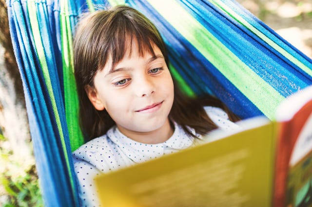 A young girl with brown hair relaxes in a colorful hammock, smiling as she reads a book outdoors. Su...
