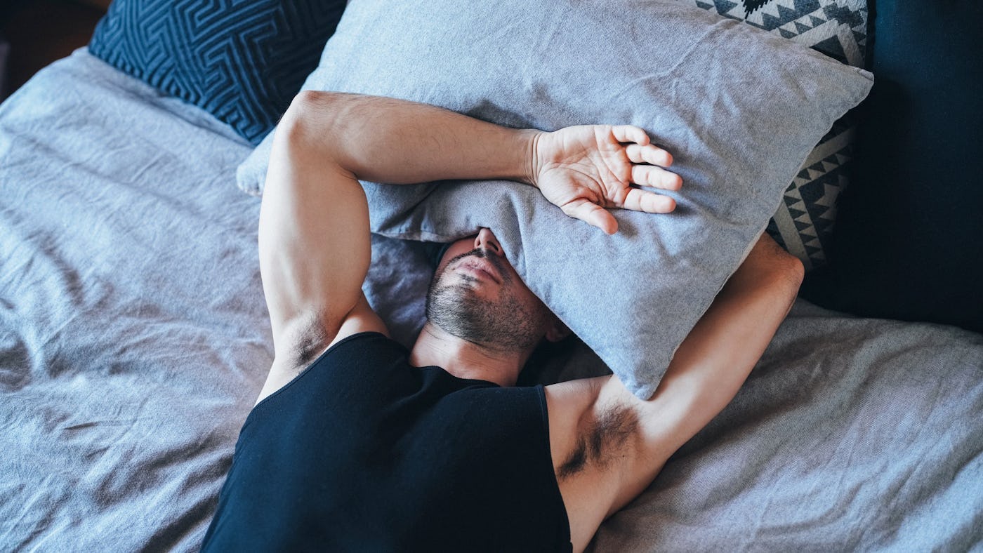 A man lies in bed, partially covered by a grey pillow, with his arms stretched out. He's wearing a sleeveless black shirt and appears to be resting.