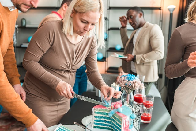 A woman in a brown dress cuts a blue and white cake at a celebration, while people socialize in the ...