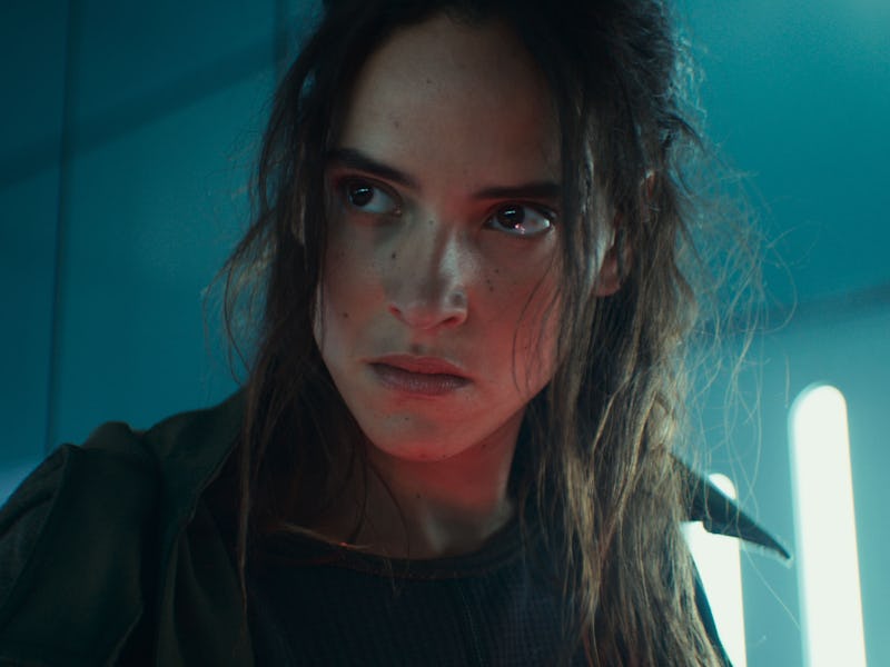 A young woman with tousled hair and an intense expression stands in a futuristic setting, illuminate...