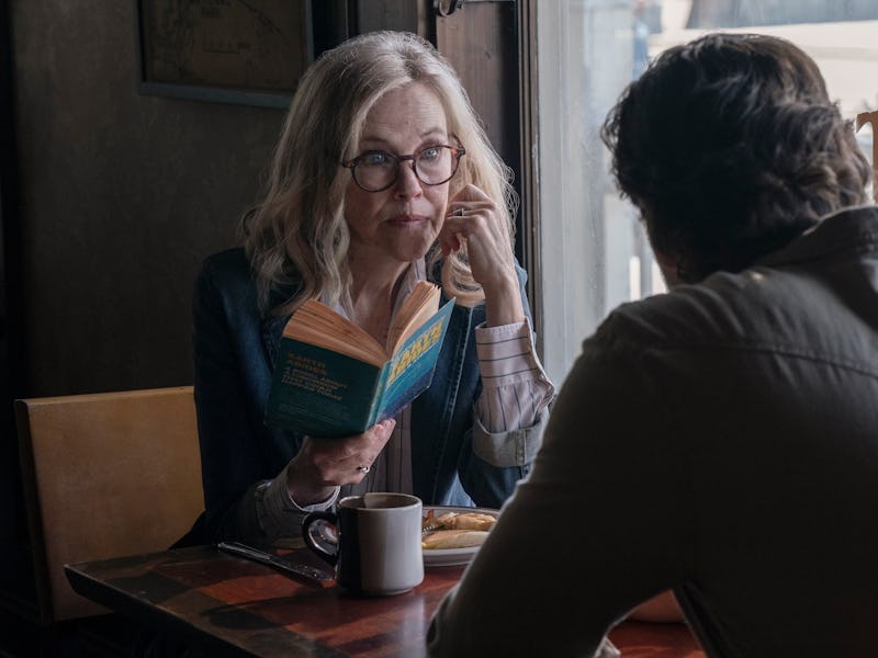 An older woman with glasses sits at a café table, reading a book while looking thoughtfully at a man...