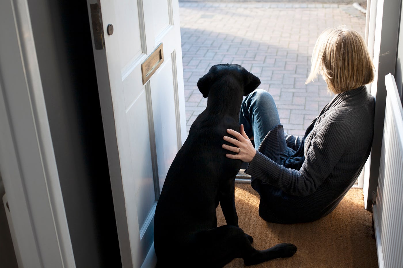 A person with short blonde hair sits by an open door on a mat, gently petting a black dog beside them, enjoying a moment of calm outdoors.