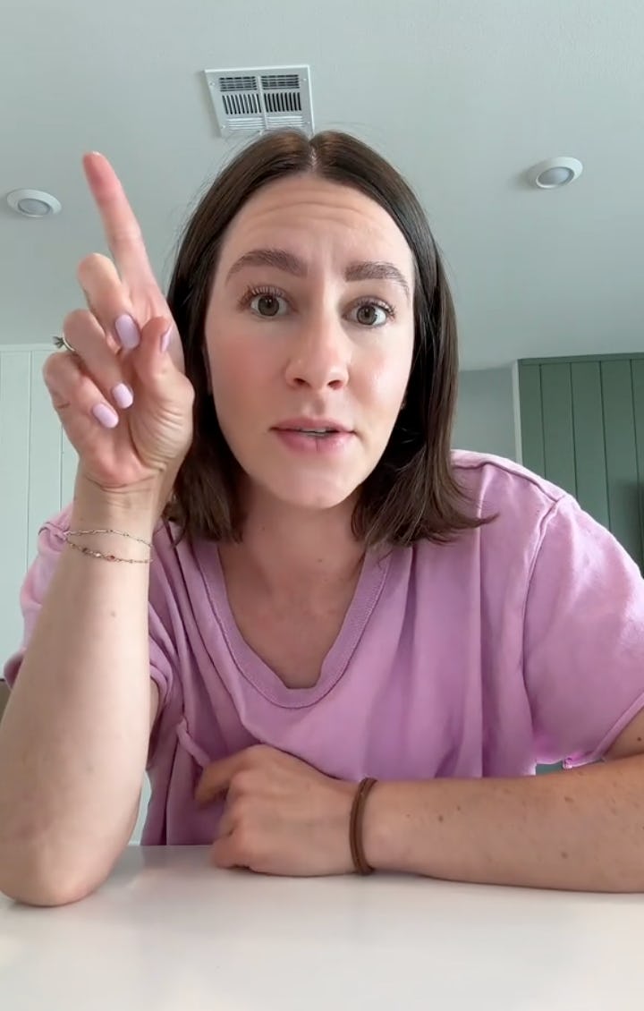 A woman with shoulder-length brown hair wears a pink shirt and gestures with her finger raised, appearing to make a point in a bright room.
