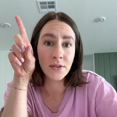 A woman with shoulder-length brown hair wears a pink shirt and gestures with her finger raised, appearing to make a point in a bright room.
