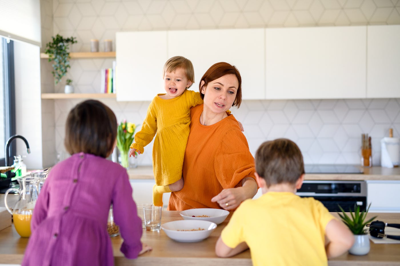 A mother in a kitchen prepares food with three children. One child is being held, while two others are seated at the table, all smiling.
