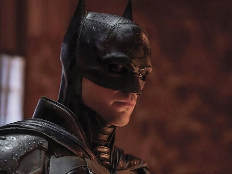 A close-up of Batman in a dark, moody setting. He wears a detailed black suit and mask, with droplet...
