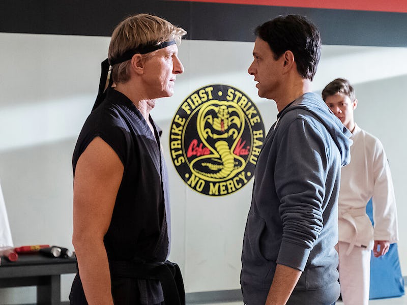 Two men face off in a dojo, tension visible in their expressions. In the background, two younger pra...