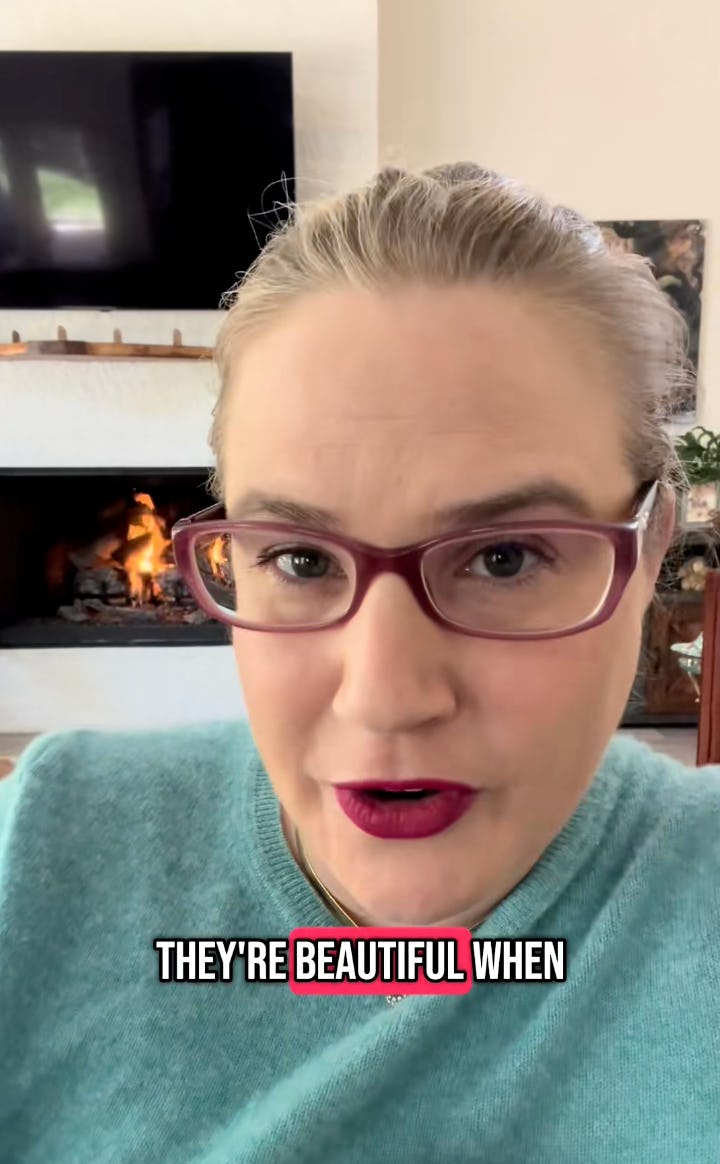 A woman with glasses and a bright lipstick smiles at the camera, sitting in a cozy room with a fireplace and a TV in the background.