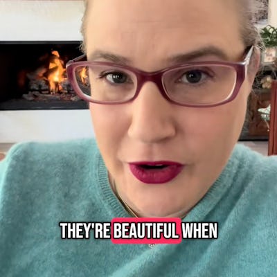 A woman with glasses and a bright lipstick smiles at the camera, sitting in a cozy room with a fireplace and a TV in the background.