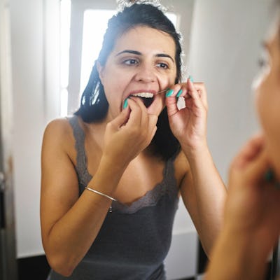 A woman with dark hair is in a bathroom, looking in the mirror while using dental floss to clean her teeth. Natural light floods the scene.