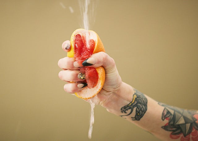 A hand with tattoos squeezes a grapefruit, causing juice to splash. The background is a soft brown, ...