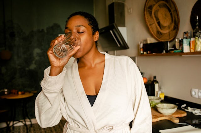 A woman in a beige robe drinks liquid collagen from a glass in a cozy kitchen, surrounded by various...