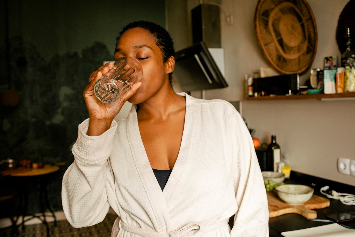 A woman in a beige robe drinks liquid collagen from a glass in a cozy kitchen, surrounded by various kitchen items and plants. Natural light illuminates the scene.