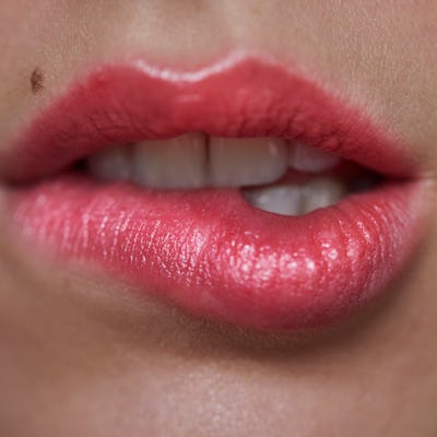 Close-up of a pair of full, gently parted lips, enhanced with glossy pink lipstick. A small mole is noticeable on the left side.