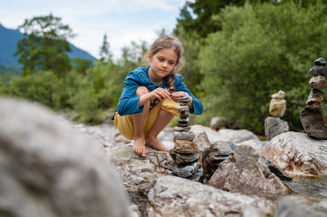 A young girl crouches on a rocky riverbank, carefully balancing stones to create a small rock tower,...