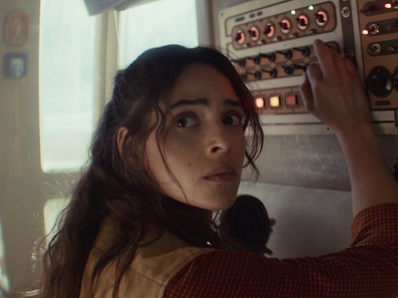 A woman with long hair looks anxiously at a control panel filled with buttons and switches, her hand...