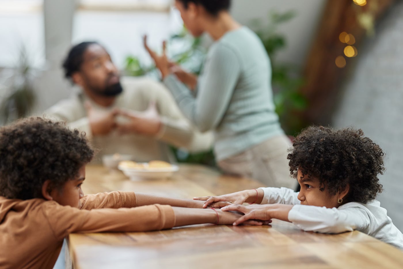 A family scene at a wooden table where two children with curly hair reach for each other. In the background, an adult gestures while communicating.