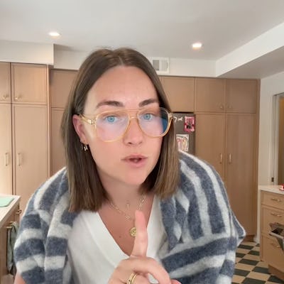 A person with medium-length brown hair, wearing glasses and a striped shawl, gestures with one finger while speaking. The kitchen features light wooden cabinets.