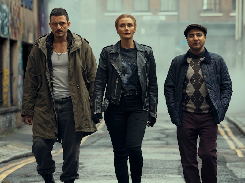 Three individuals walk confidently down a foggy, urban street. The man on the left wears a long coat...