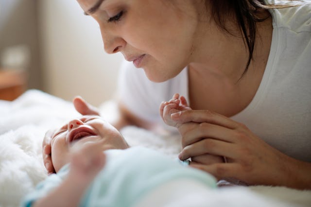 A mother gently holds her baby’s head while smiling, as the baby lies on a soft surface, reaching ou...